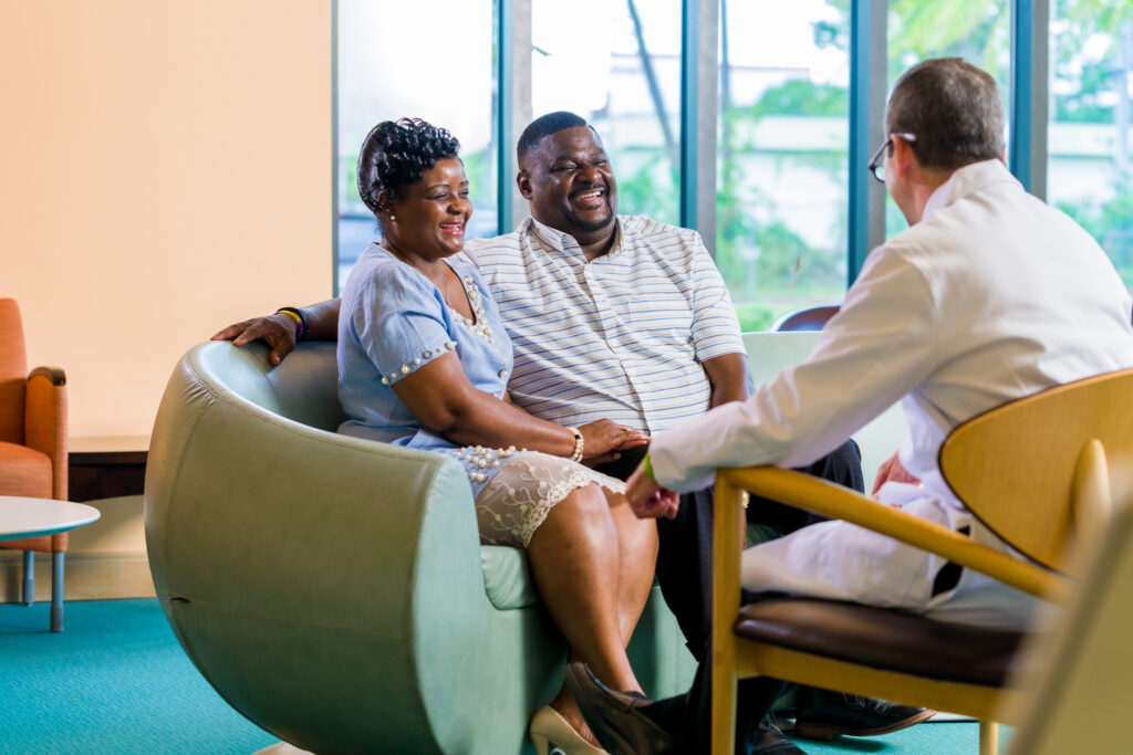 A smiling middle-aged couple sits closely together on a light green curved sofa, engaged in conversation with a man in a white coat seated on a wooden chair facing them. The woman wears a light blue dress with pearl embellishments and beige heels, while the man wears a white and gray striped shirt and black pants. The setting appears to be a bright, modern room with large windows and teal carpeting.