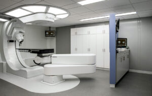 A modern medical treatment room featuring a large white robotic radiation therapy machine labeled "RoboCouch" with an adjustable patient table. The room has white cabinets along the back wall, a blue partition with mounted monitors displaying medical imaging, and a clean, clinical design with gray flooring and a white ceiling with recessed lighting.