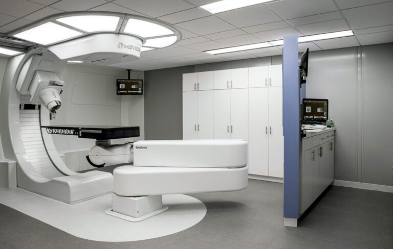 A modern medical treatment room featuring a large white robotic radiation therapy machine labeled "RoboCouch" with an adjustable patient table. The room has white cabinets along the back wall, a blue partition with mounted monitors displaying medical imaging, and a clean, clinical design with gray flooring and a white ceiling with recessed lighting.