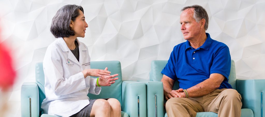 A female doctor in a white coat is sitting and talking with an older man wearing a blue polo shirt and beige pants. They are seated on light blue chairs against a white textured wall. The doctor appears to be explaining something with hand gestures while the man listens attentively.