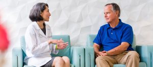 A female doctor in a white coat is sitting and talking with an older man wearing a blue polo shirt and beige pants. They are seated on light blue chairs against a white textured wall. The doctor appears to be explaining something with hand gestures while the man listens attentively.