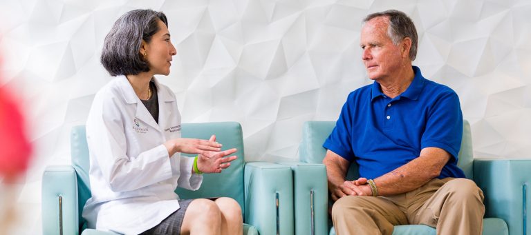 A female doctor in a white coat is sitting and talking with an older man wearing a blue polo shirt and beige pants. They are seated on light blue chairs against a white textured wall. The doctor appears to be explaining something with hand gestures while the man listens attentively.