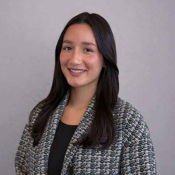 Young woman with long dark hair wearing a patterned jacket over a black top, smiling against a plain light gray background.