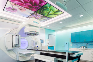 A modern radiation therapy room featuring an Elekta Versa HD linear accelerator machine. The room has a sleek design with light blue walls, blue cabinets, and a tiled backsplash. The ceiling is decorated with illuminated panels displaying large, colorful flower images. The treatment table is black with white and blue components, and there is a control panel attached to it. The room is brightly lit with recessed lighting.