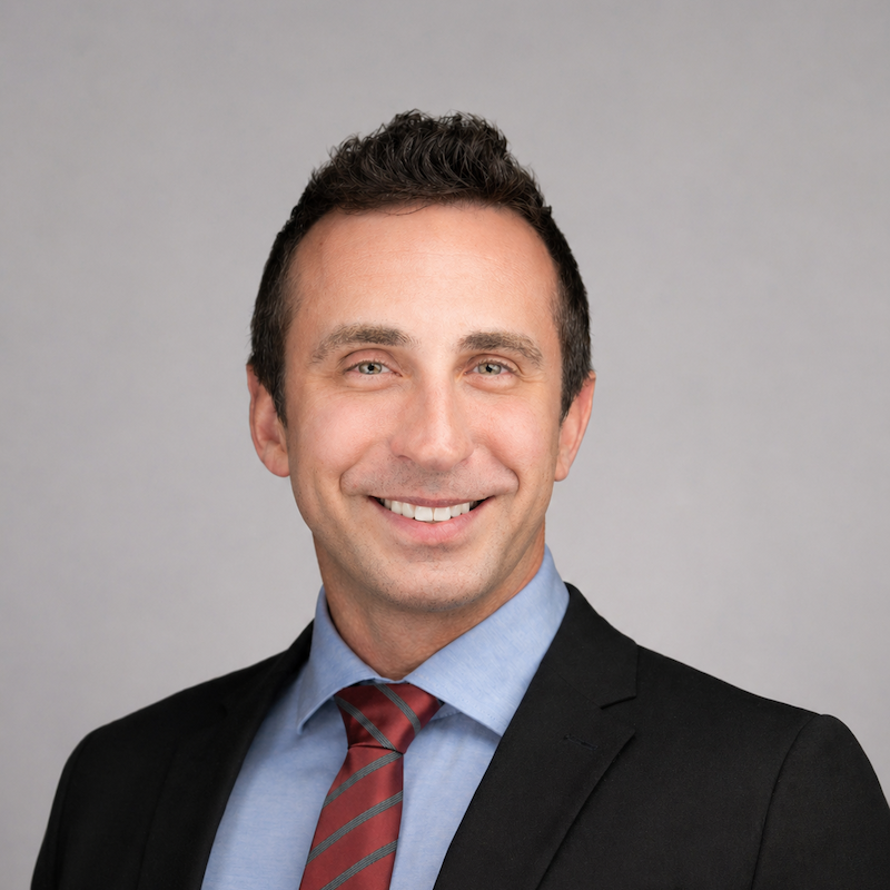 Man with short dark hair wearing a black suit jacket, light blue dress shirt, and red striped tie, smiling against a plain light gray background.